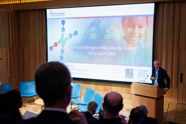 Oxford-Harrington Director Matthew Wood speaks at a lectern to the symposium audience at Trinity College, Oxford. A presentation screen next to him has the text 
